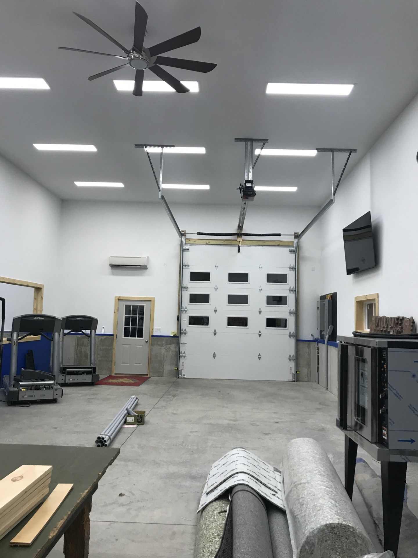 An indoor garage or workspace with white walls, a large roll-up door, a ceiling fan, LED lighting, and fitness equipment.