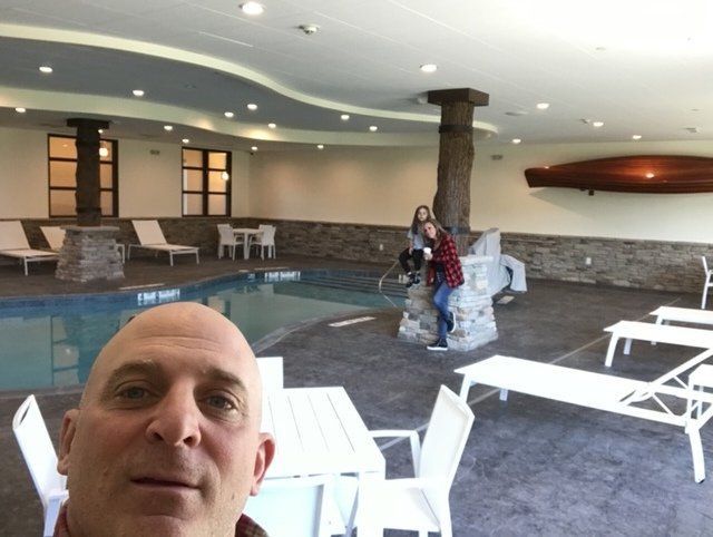 Man takes a selfie by an indoor pool with lounge chairs, tables, and two people near a tree pillar.