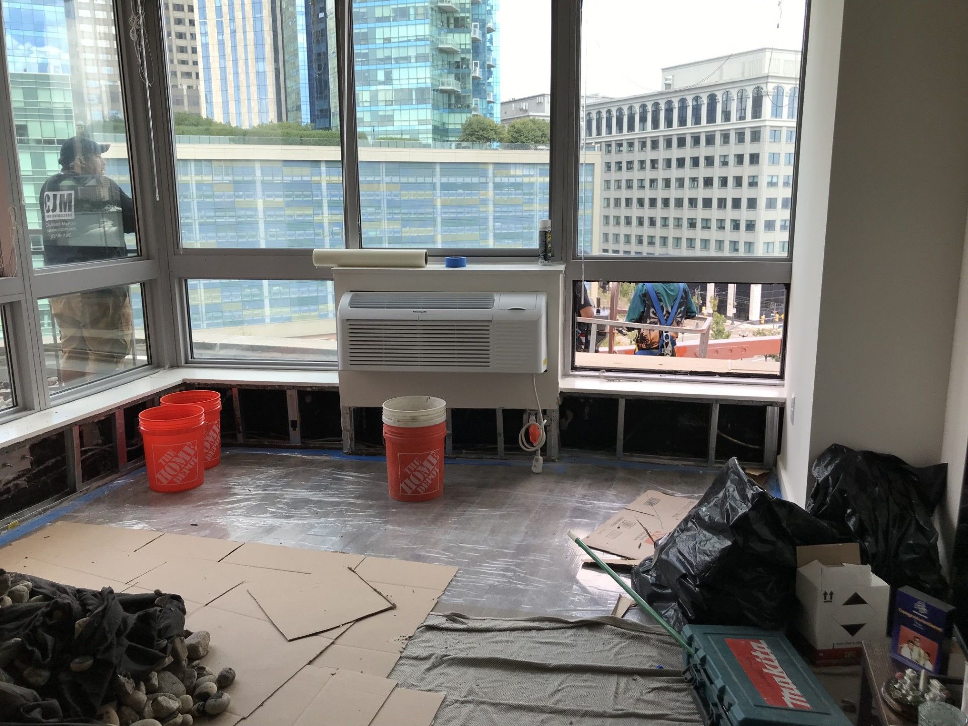 A room under construction with an HVAC unit below windows, orange buckets, floor protection, and bags of debris.