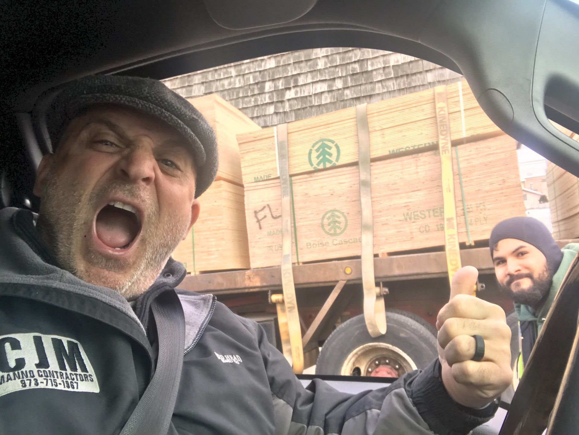 Two people in a vehicle, one in the foreground yelling with a thumbs-up, and a flatbed truck carrying wood in the back.