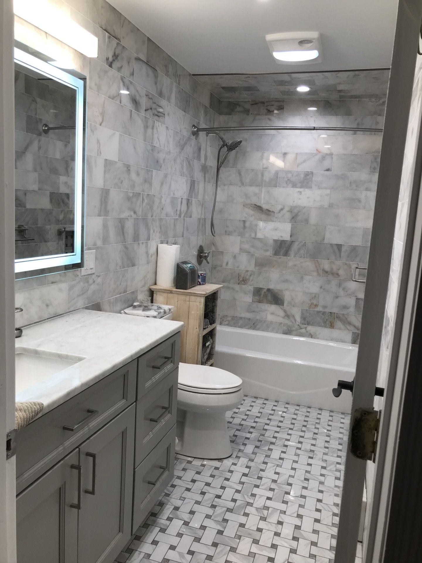 A gray vanity with a marble countertop, an LED-lit mirror, a toilet, and a bathtub with marble-tiled walls in a bathroom.