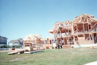House under construction, wooden frame, on a green lawn with other houses visible in the background under a blue sky.