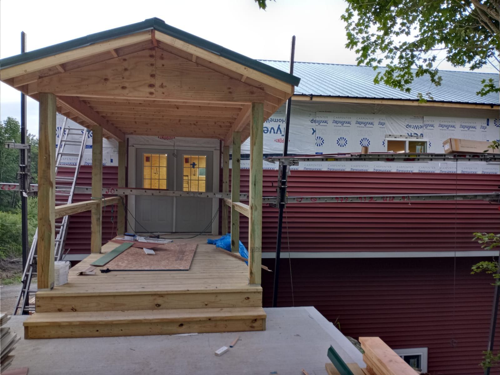 A wooden porch structure under construction in front of a building with red metal siding and white exterior sheathing.