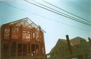 Wooden house frame under construction next to a finished house on a sunny day. Power lines overhead.