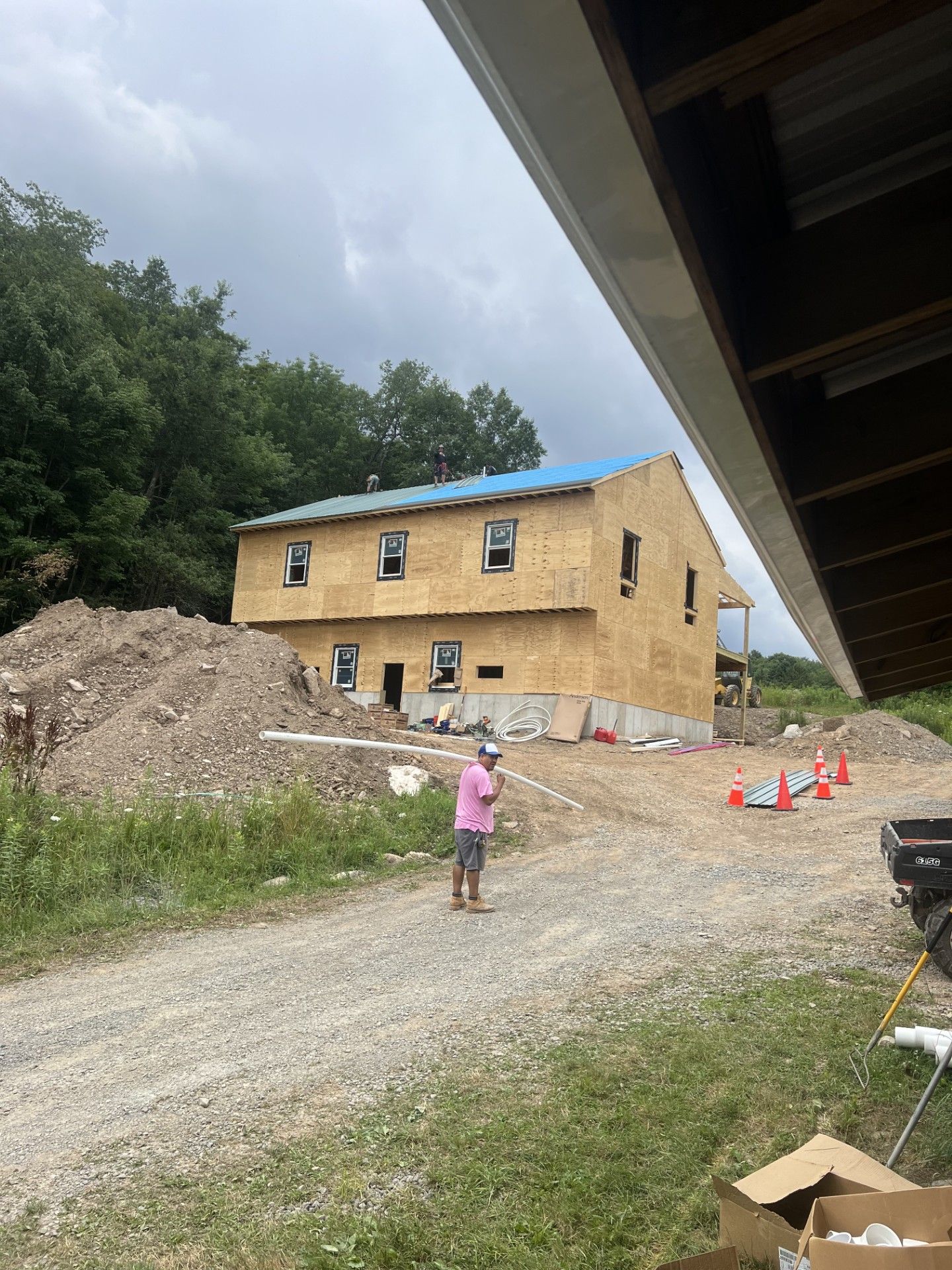 A two-story house under construction with wood sheathing and a blue roof, viewed from a gravel construction site.