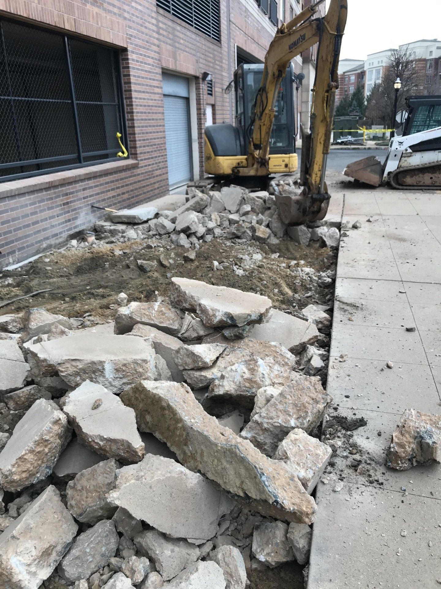 A yellow excavator is parked beside a building, breaking up concrete slabs along a sidewalk.