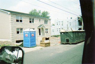 A light brick apartment building under construction, with a blue porta-potty and a green dumpster in front.