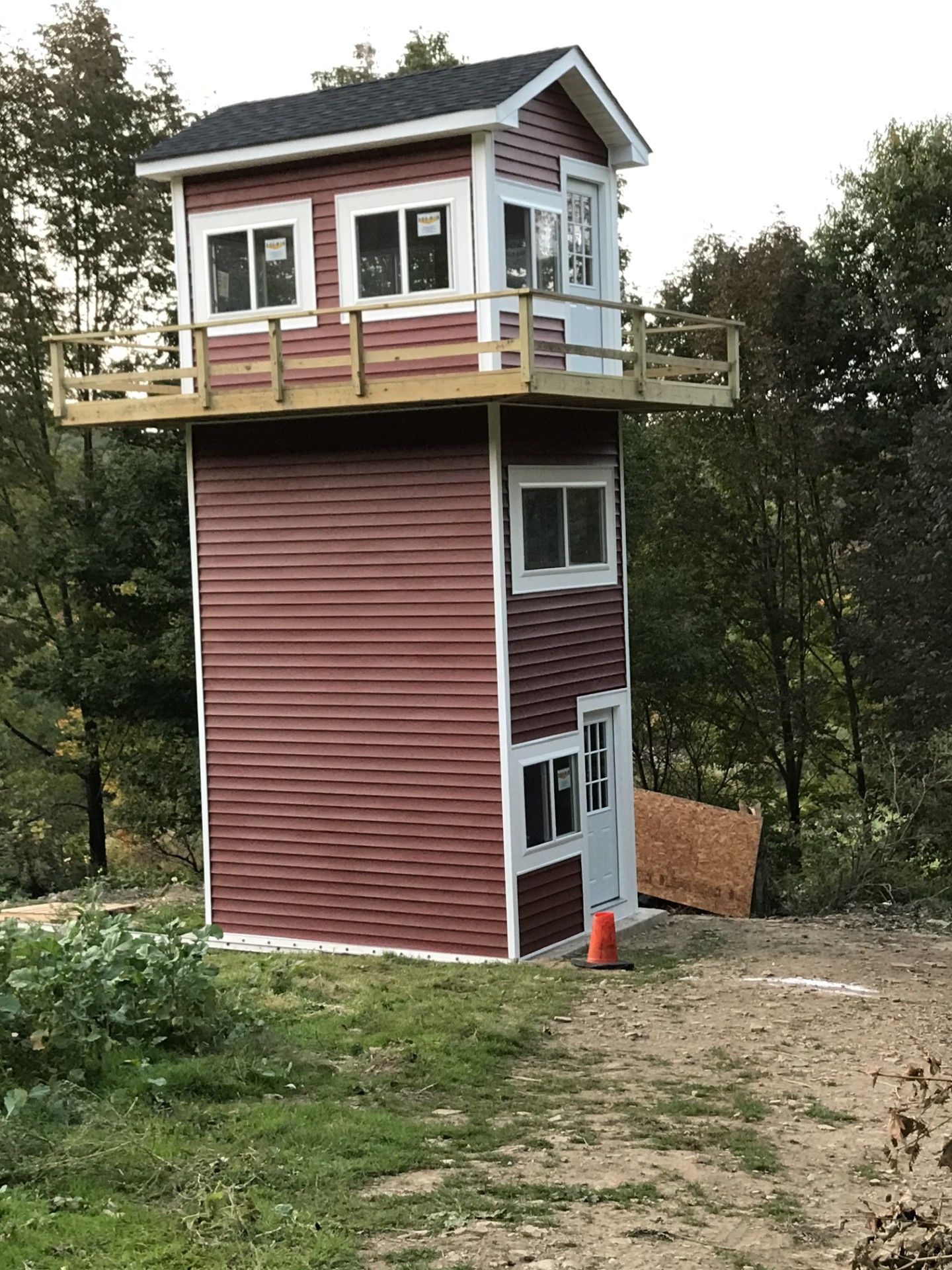 A two-story, red-sided tower with white trim, a surrounding deck on the top level, and windows, set in a wooded area.