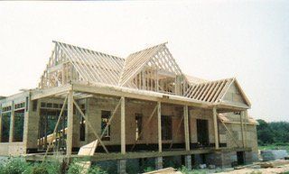 House under construction, wooden frame, roof trusses in place, porch, daylight setting.