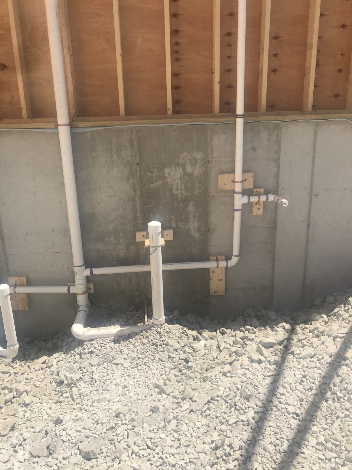 White PVC plumbing pipes attached to a concrete foundation wall in an unfinished room with gravel flooring.