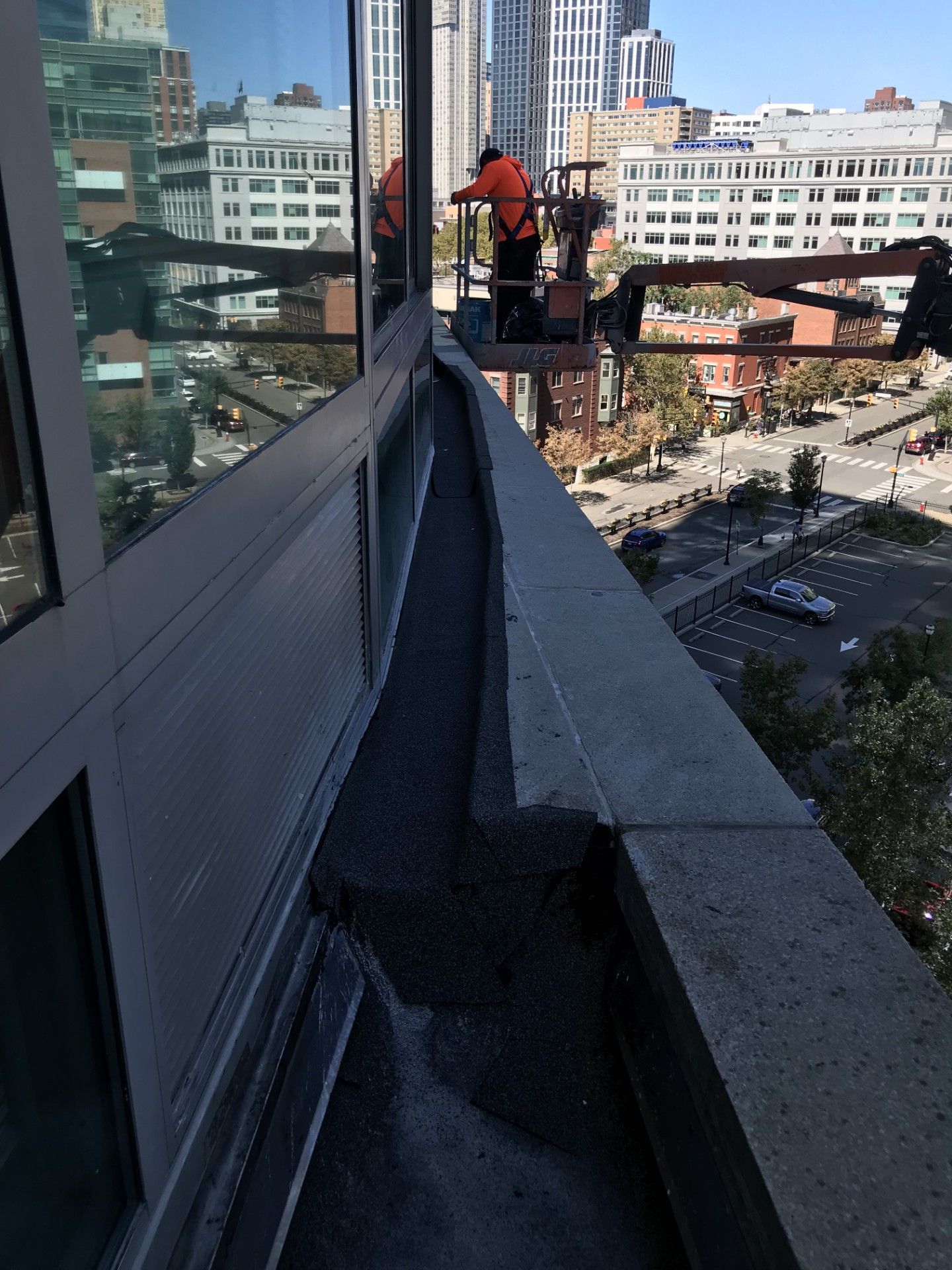 A construction worker in an orange vest stands in a lift basket against the glass exterior of a high-rise building.