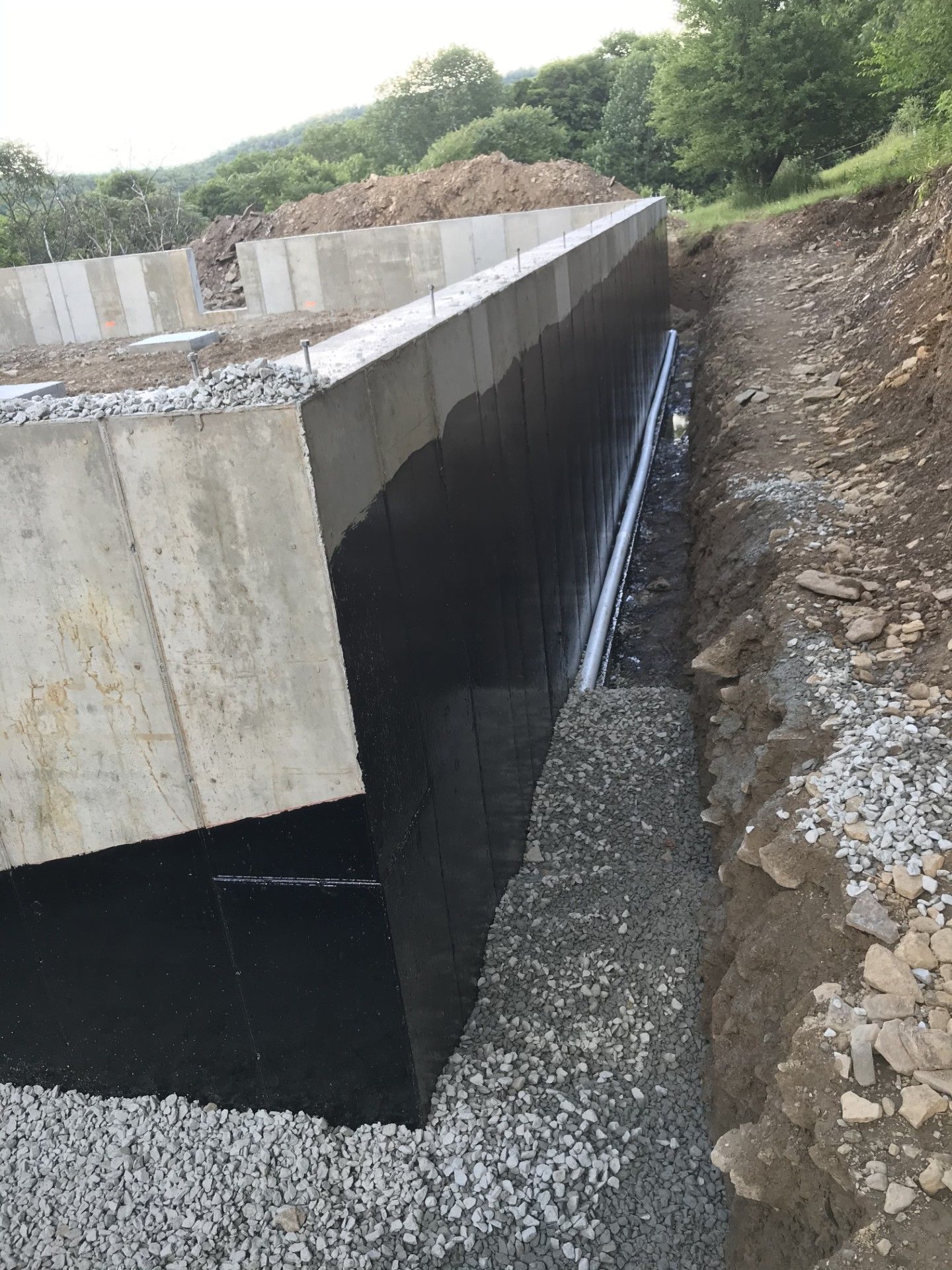 A concrete foundation wall with black waterproofing, a drainage pipe, and gravel base along an excavated dirt slope.