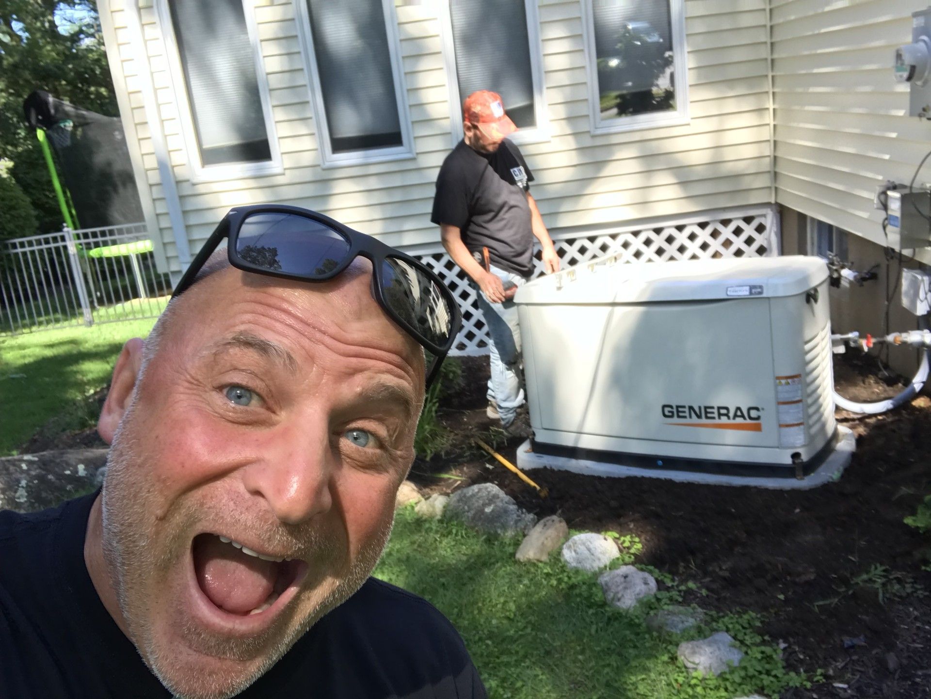 A man smiles excitedly in the foreground while a worker stands near a Generac backup generator outside a house.