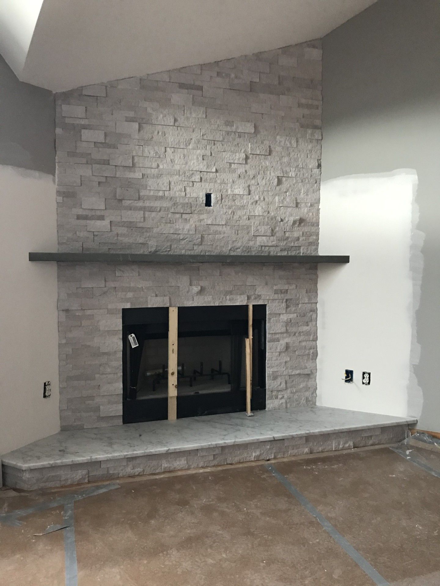 An unfinished fireplace with gray stacked stone, a dark mantel, and a black insert, centered on a light-colored wall.