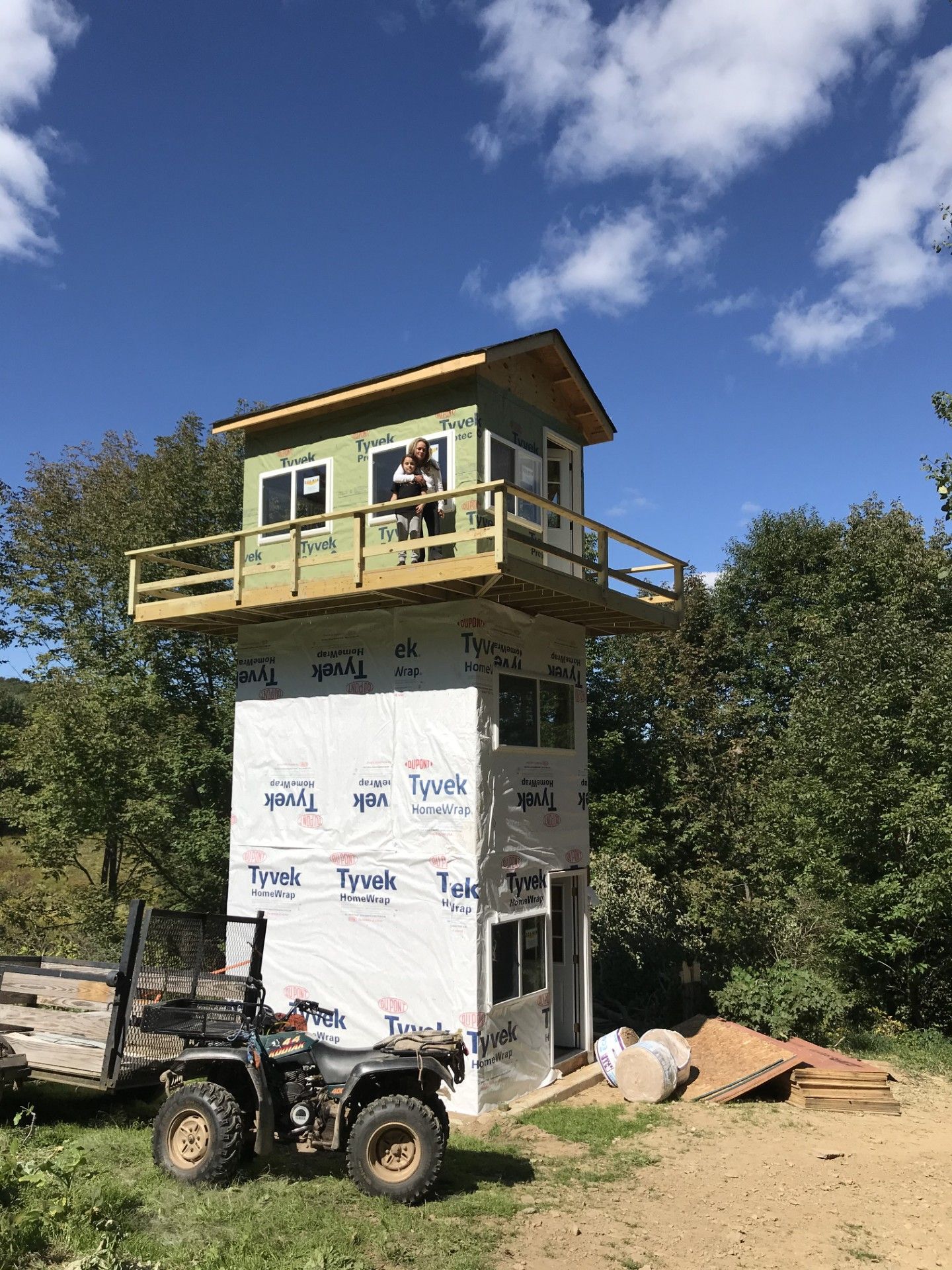 A two-story tower under construction in a wooded area with an ATV parked in front and a person standing on the balcony.