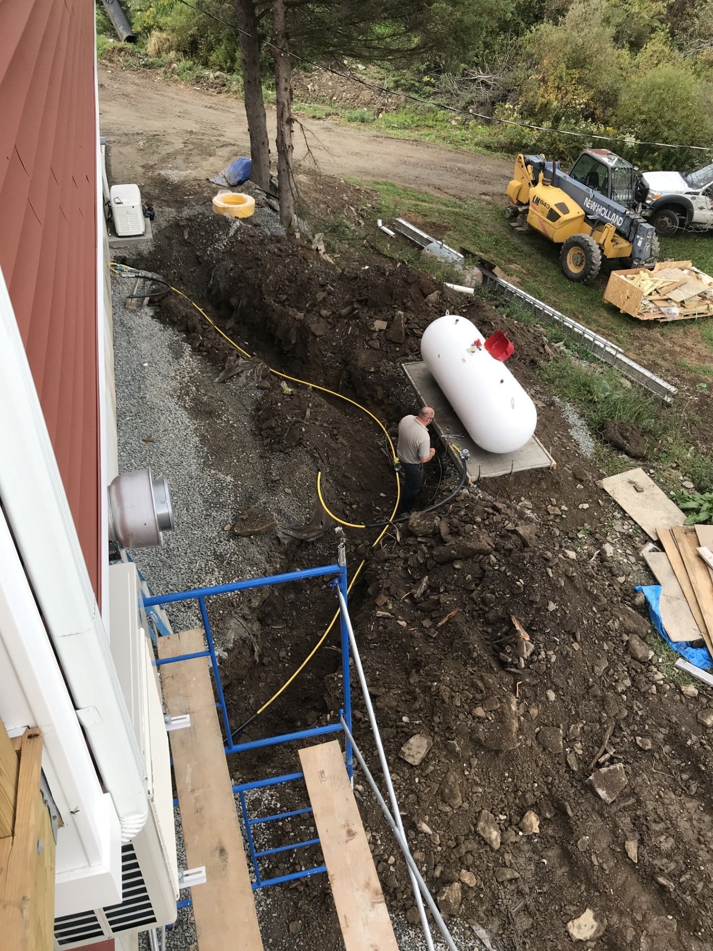 A worker installing a yellow gas line in a trench next to a house and a large propane tank.