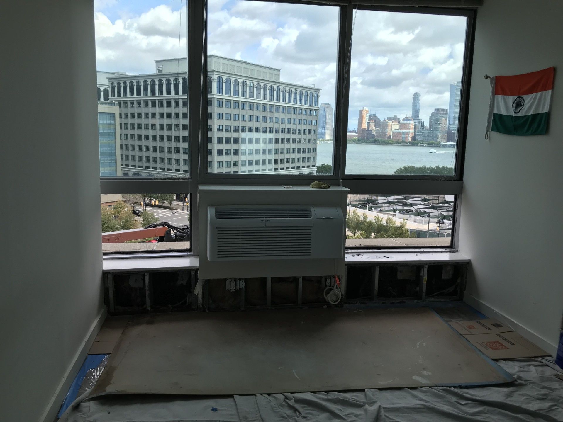 A room view showing large windows overlooking a city and body of water, with an air conditioner and an Indian flag.