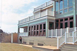 Two-story house with green walls, brown windows, and white railings. A person stands on a wooden deck.