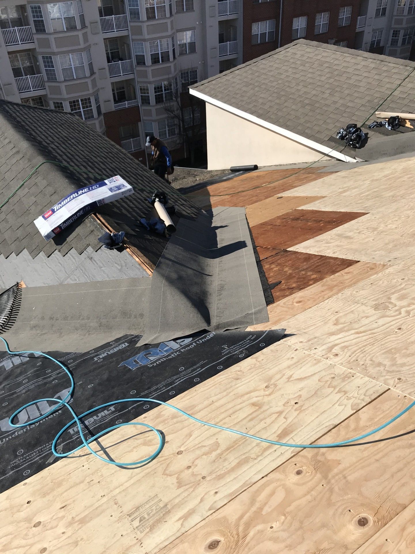A construction worker walks across a residential roof under repair, showing exposed wood decking and layers of underlayment.