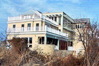 Multi-story, beige building with white trim, balconies, and a glass-enclosed section; set among dry, brushy plants.
