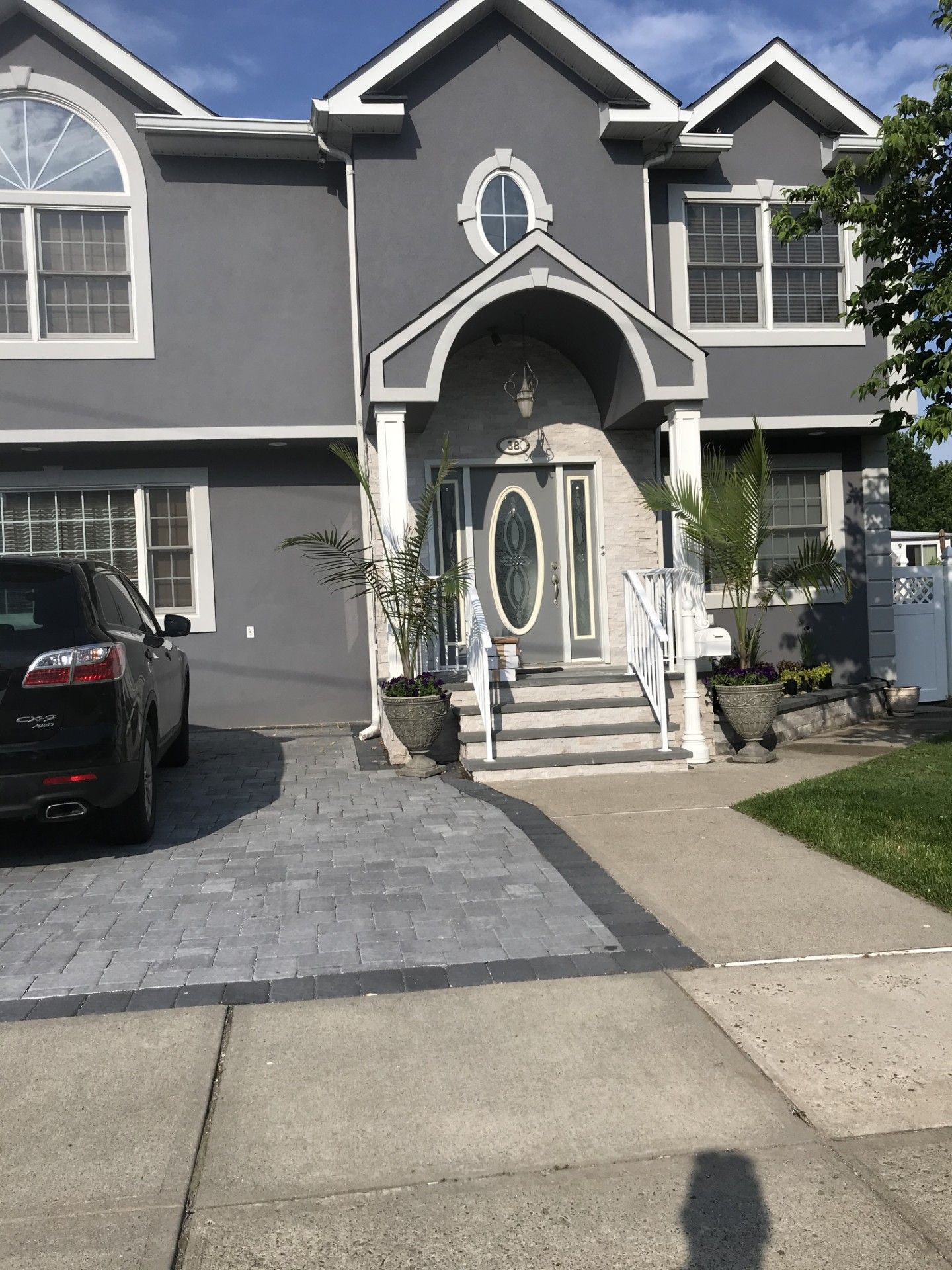 A two-story, gray house with a peaked roof, arched entryway, and gray paver driveway with a black car parked to the side.