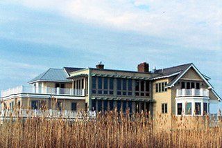 Large yellow house with multi-level decks and many windows, seen through tall brown reeds, under a blue sky.