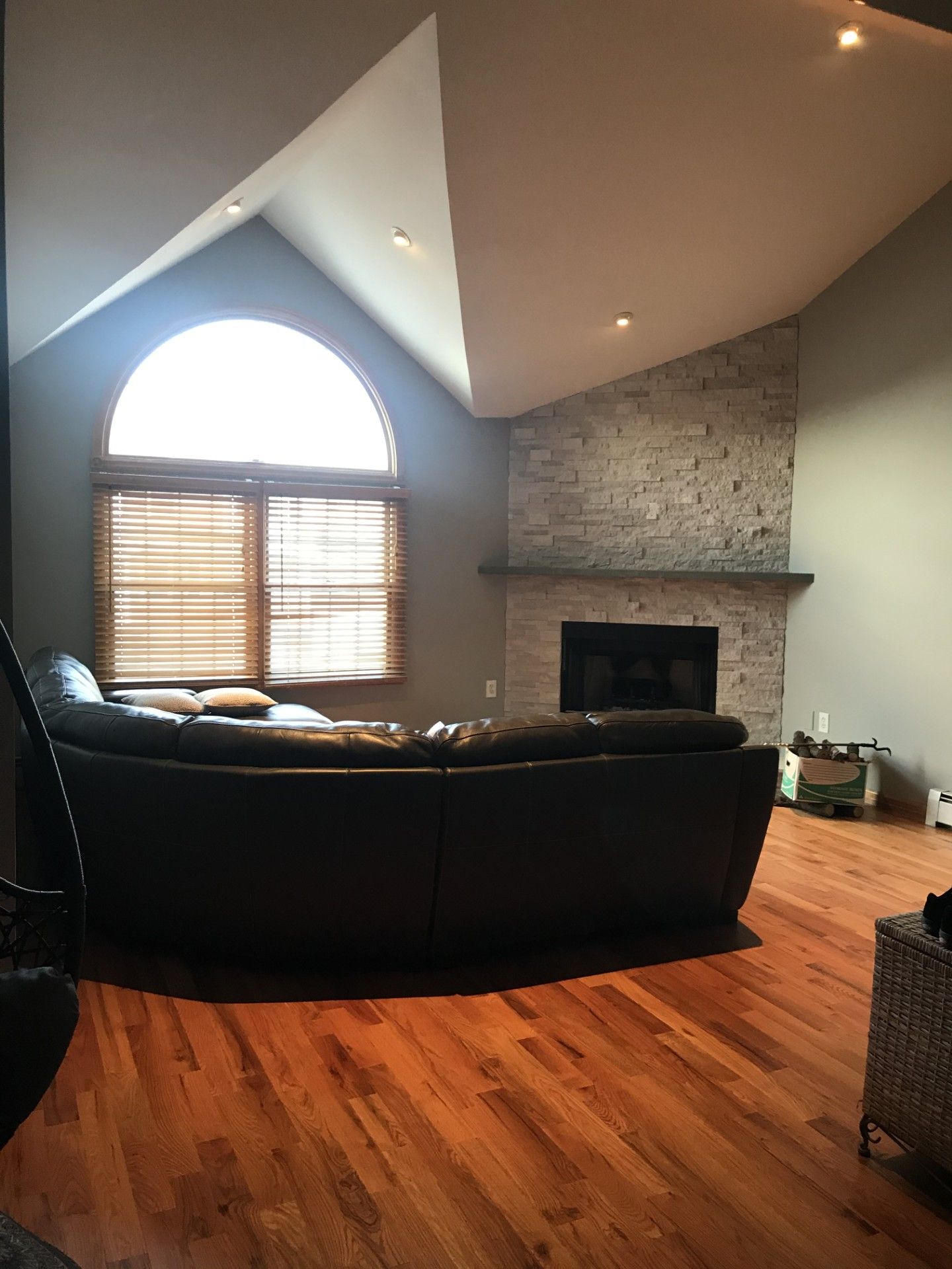 A living room with hardwood floors, a stone fireplace, and a large arched window above a curved black leather sectional.