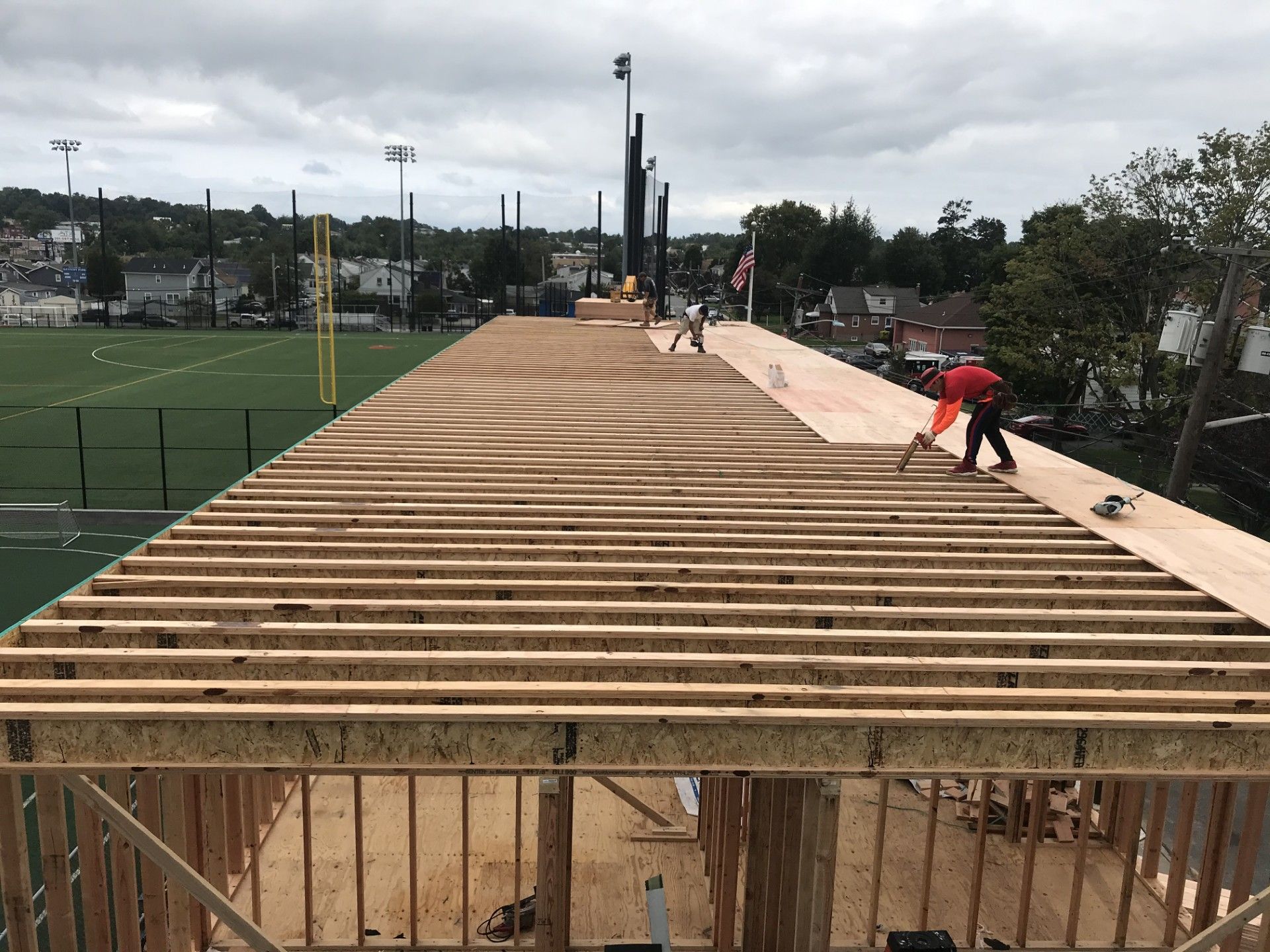 Construction workers install plywood sheathing on roof joists above an open-framed building next to a sports field.