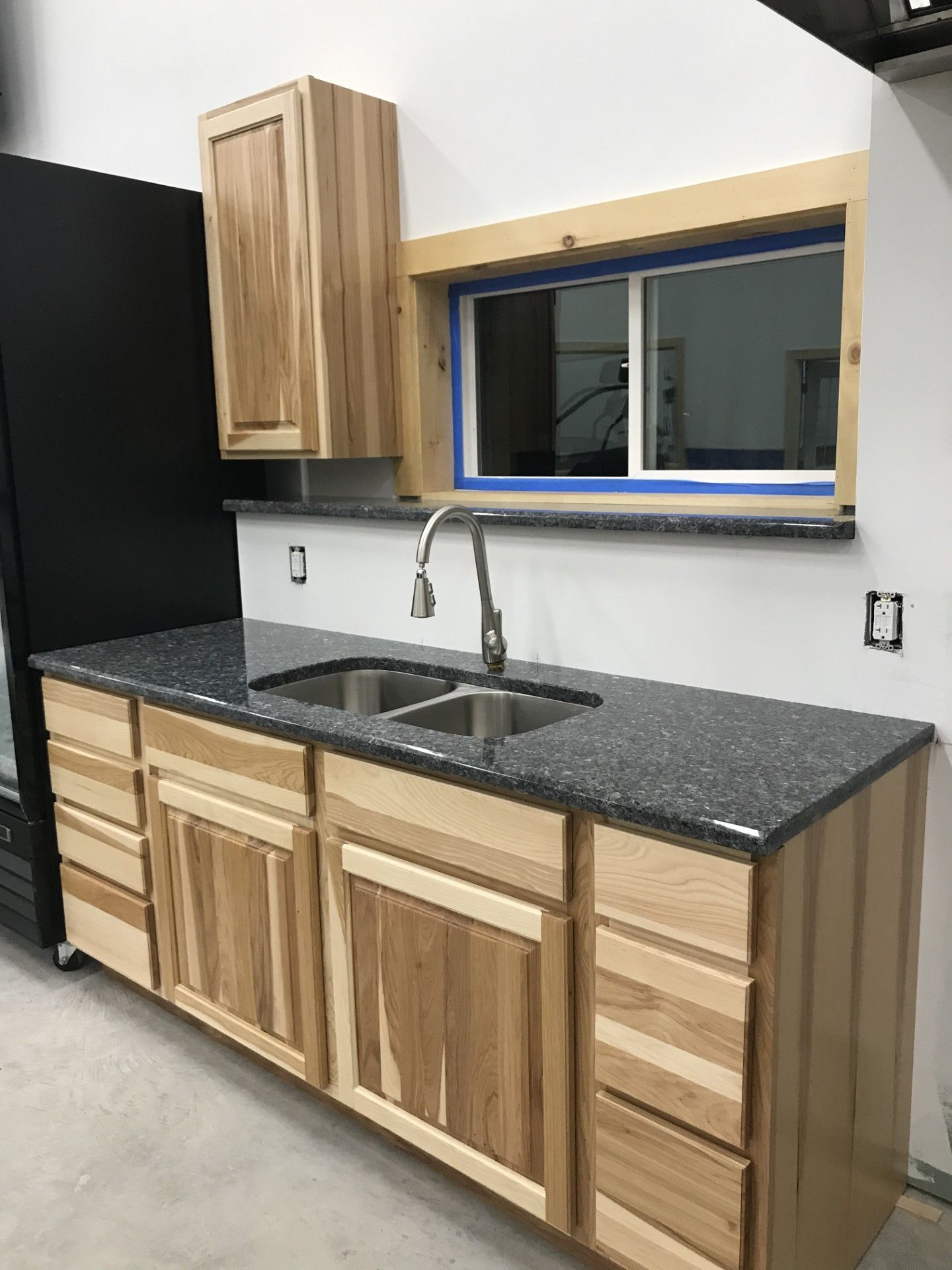 Natural wood cabinets with a dark speckled countertop and a stainless steel sink in a room with a window and black fridge.