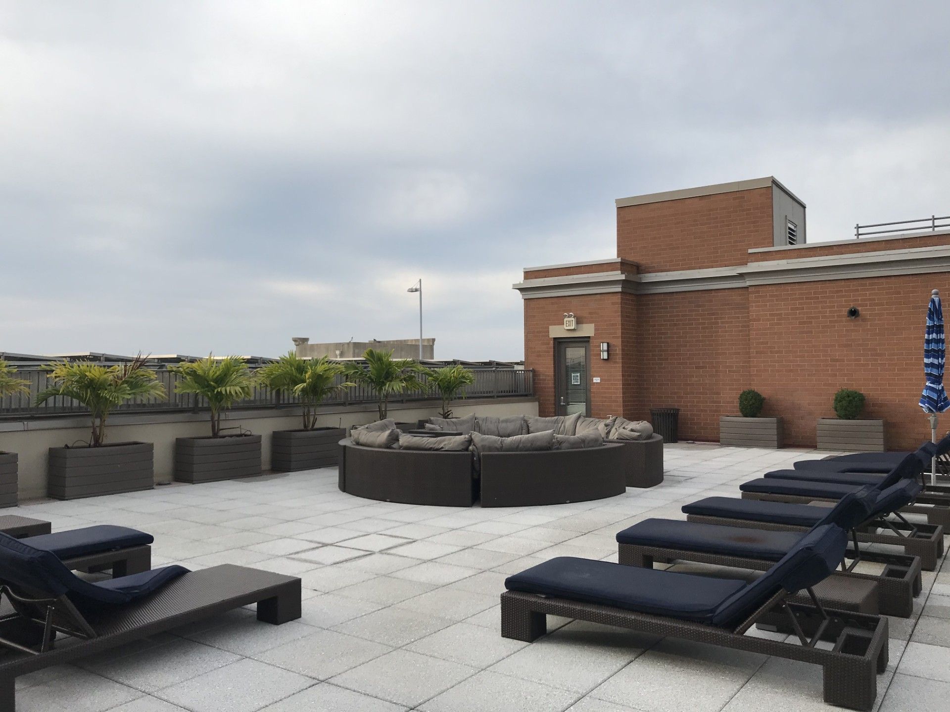Rooftop terrace with a circular wicker sofa, lounge chairs, and potted plants against a brick building on a cloudy day.