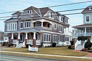 Two-story gray beach house with multiple dormers, white trim, and a porch, on a grassy lawn.