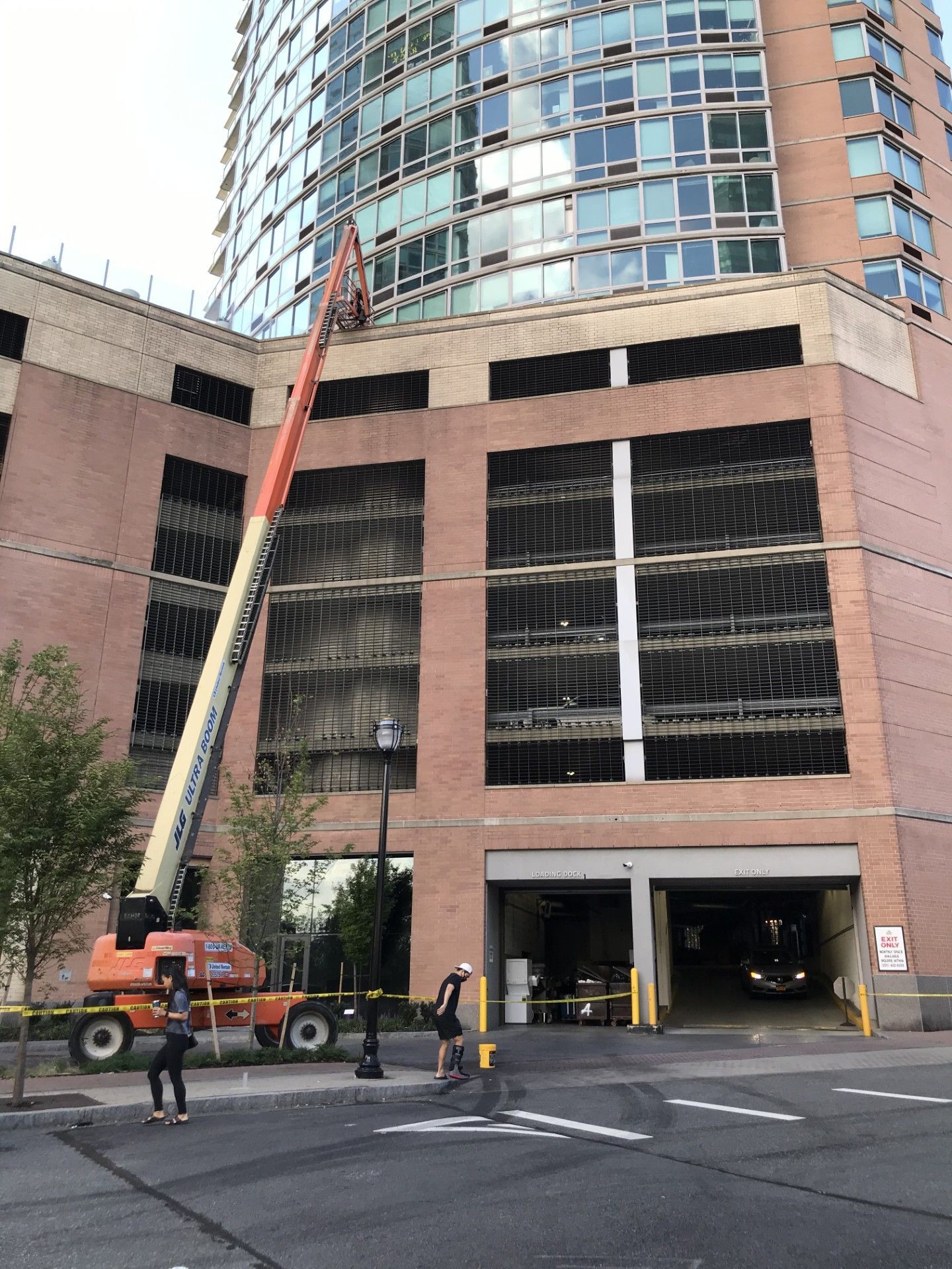 A construction lift reaches toward a tall brick building with a parking garage entrance at street level.