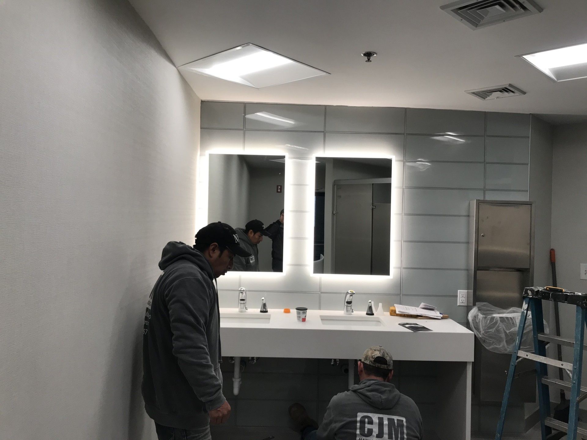 Two workers installing a modern, white bathroom vanity with backlit mirrors and gray tiled walls.