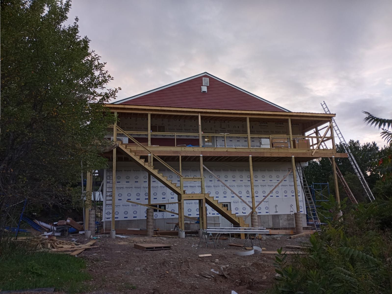A two-story house under construction with a wooden deck, staircase, white insulation, and a red-tiled roof.
