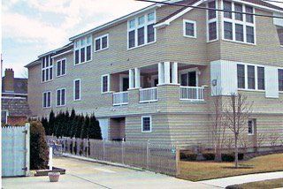 Beige multi-story building with white trim, porch, and windows; a wooden fence in the foreground.