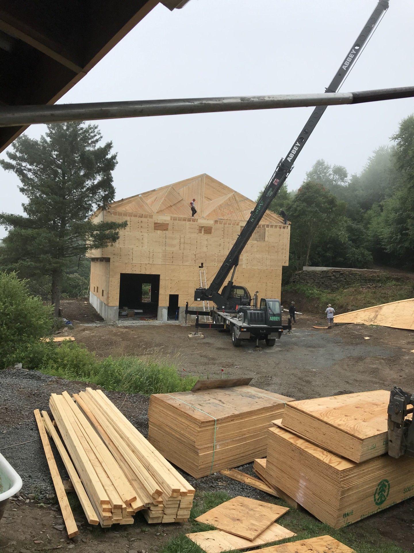 A crane lifts materials to the wooden frame of a two-story building under construction on a foggy, wooded lot.