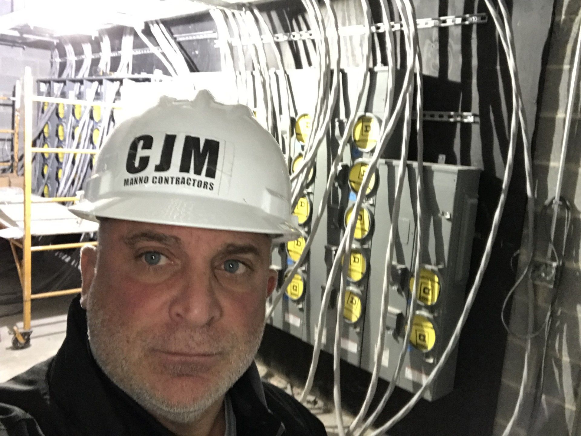 A person wearing a CJM white hard hat stands in front of a wall with multiple electrical meters and conduit piping.