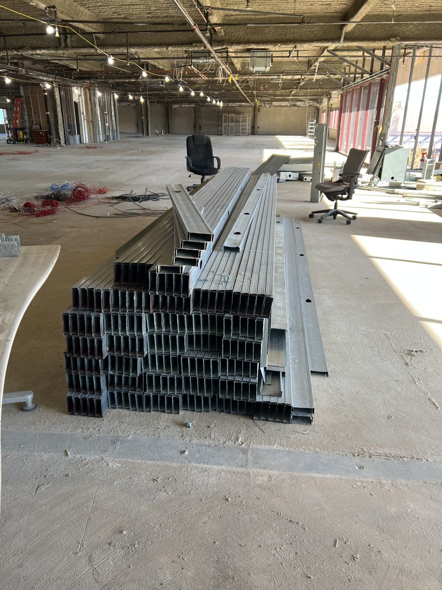 A stack of metal construction studs sits on a concrete floor in an unfinished, open-plan building with office chairs.