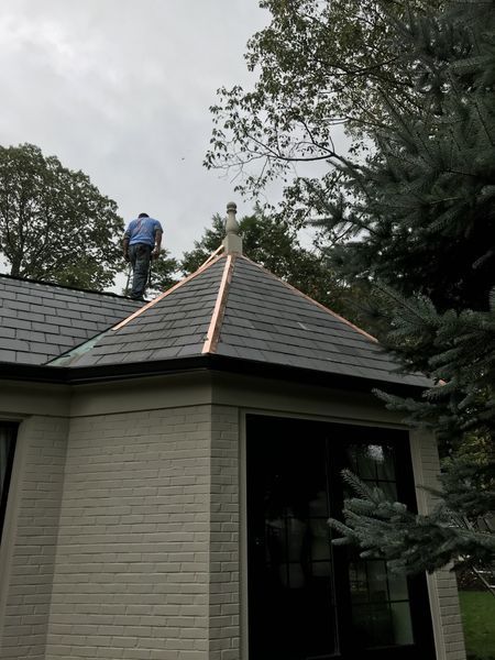 A person on a slate roof with copper flashing. Cloudy sky, trees, and a light-colored brick building are in the background.