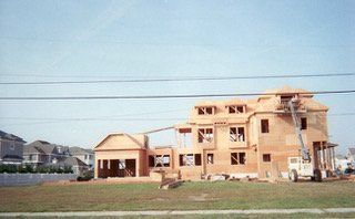 House under construction, frame visible. Construction worker on lift. Bright blue sky, sunny day.