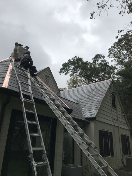 Two workers on a roof, near a chimney, with ladders. Overcast sky.