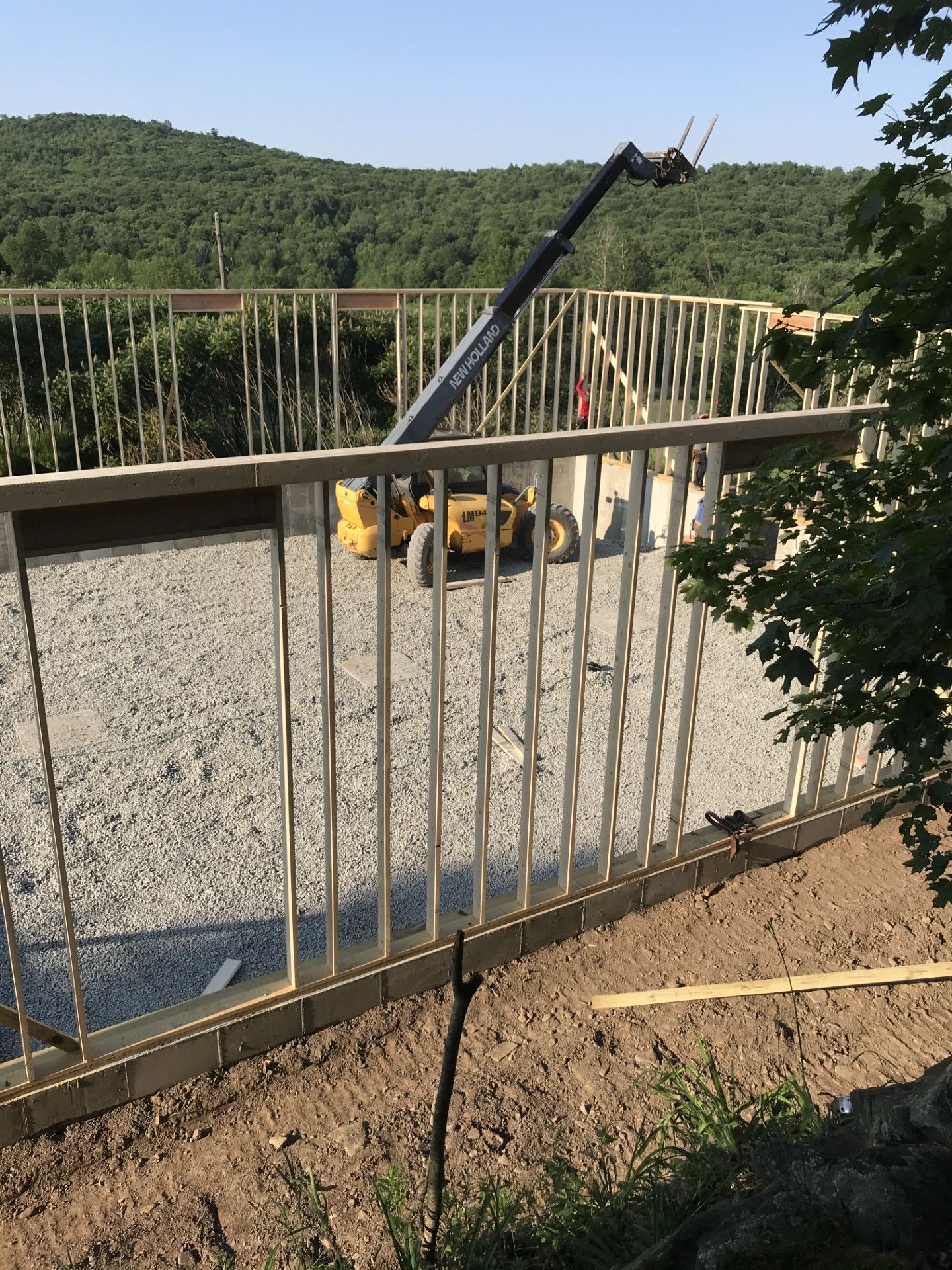 A yellow telehandler lifts materials inside a wooden frame structure built on a gravel base under a clear blue sky.