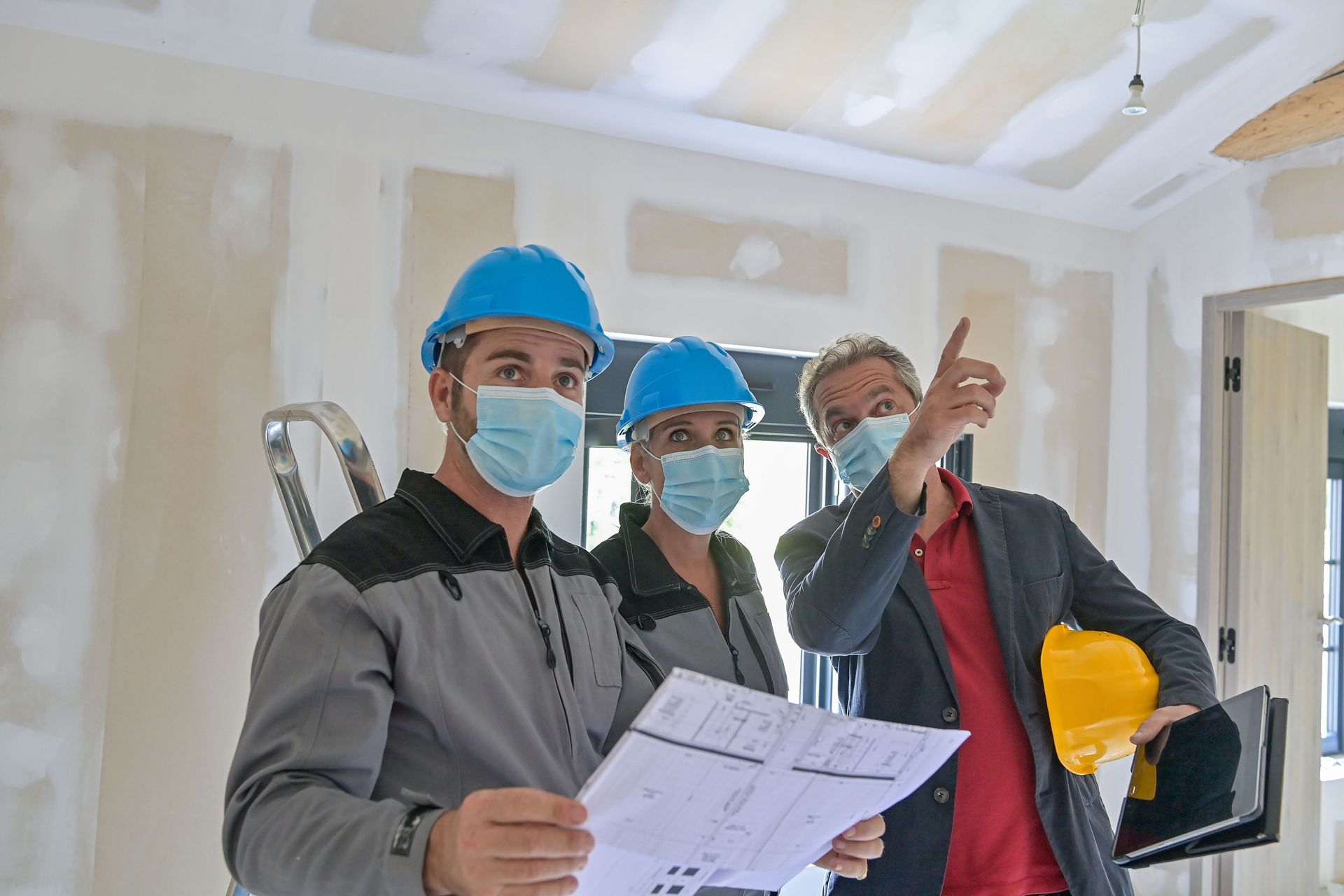 Three people in a construction site with drywall, wearing masks and helmets, reviewing blueprints and pointing at walls.