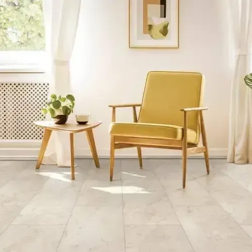 Yellow chair in a bright room with marble-look flooring, a small table with a plant, and a window with white curtains.