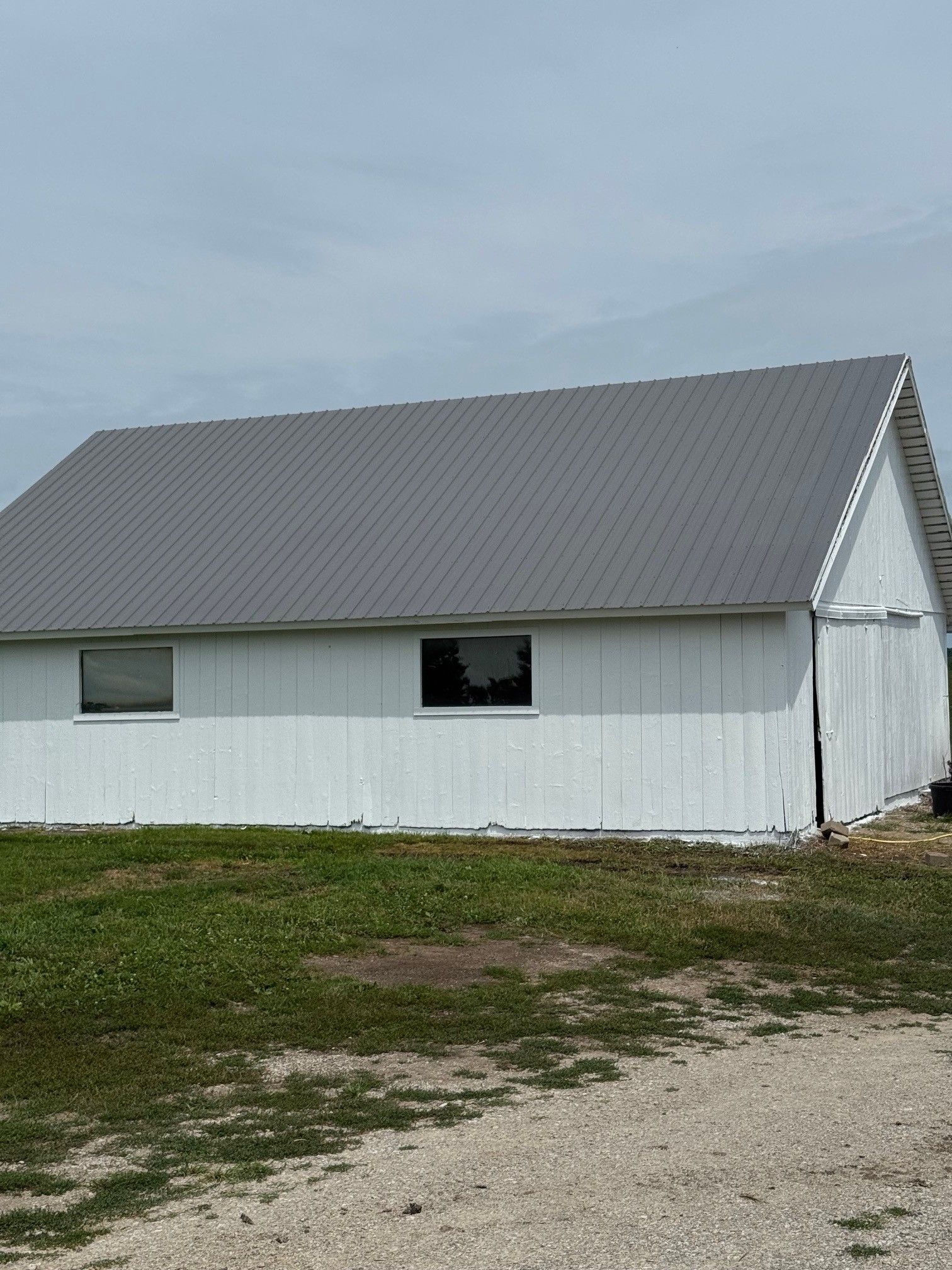 White barn with gray metal roof on a grassy dirt lot under a cloudy sky
