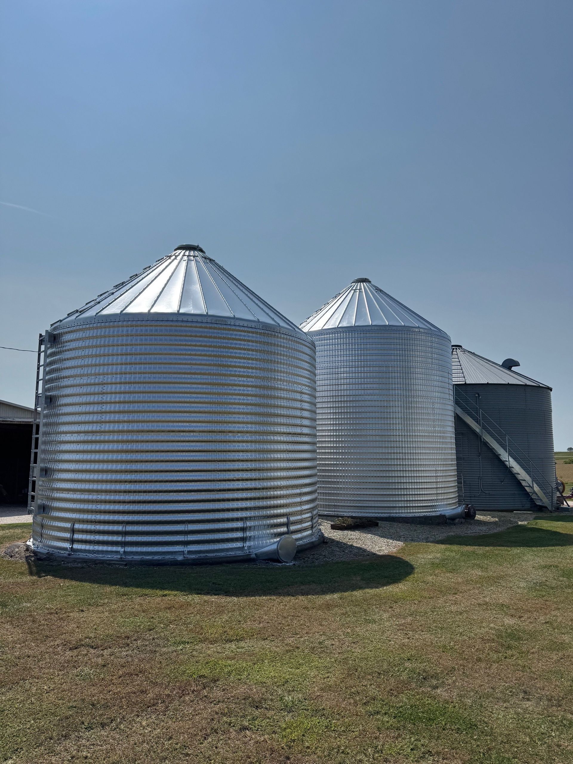 Two silver grain silos with conical roofs on a grassy farm under a clear blue sky