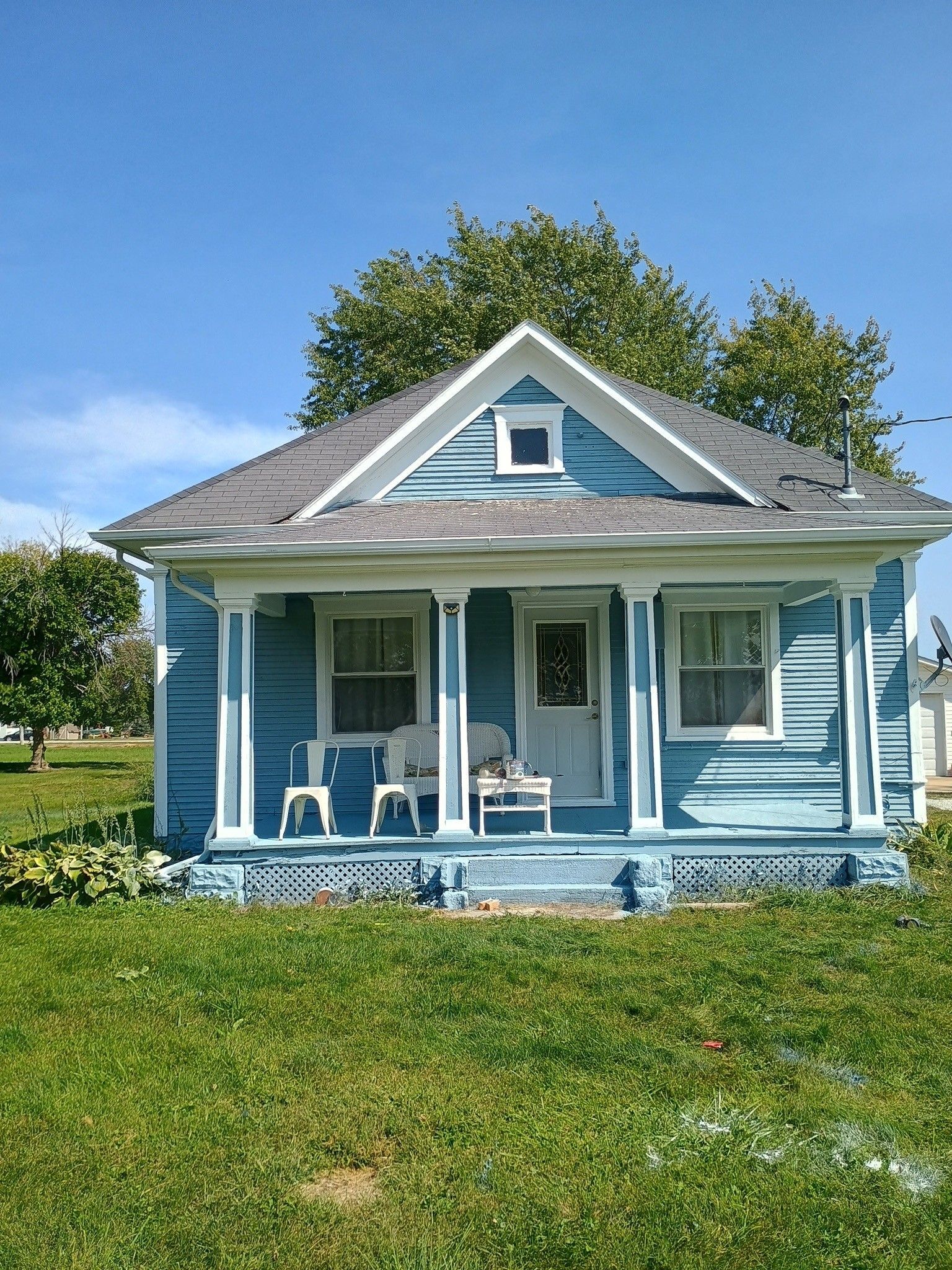 Blue cottage with a front porch and white trim, standing in a sunny grassy yard under a clear sky