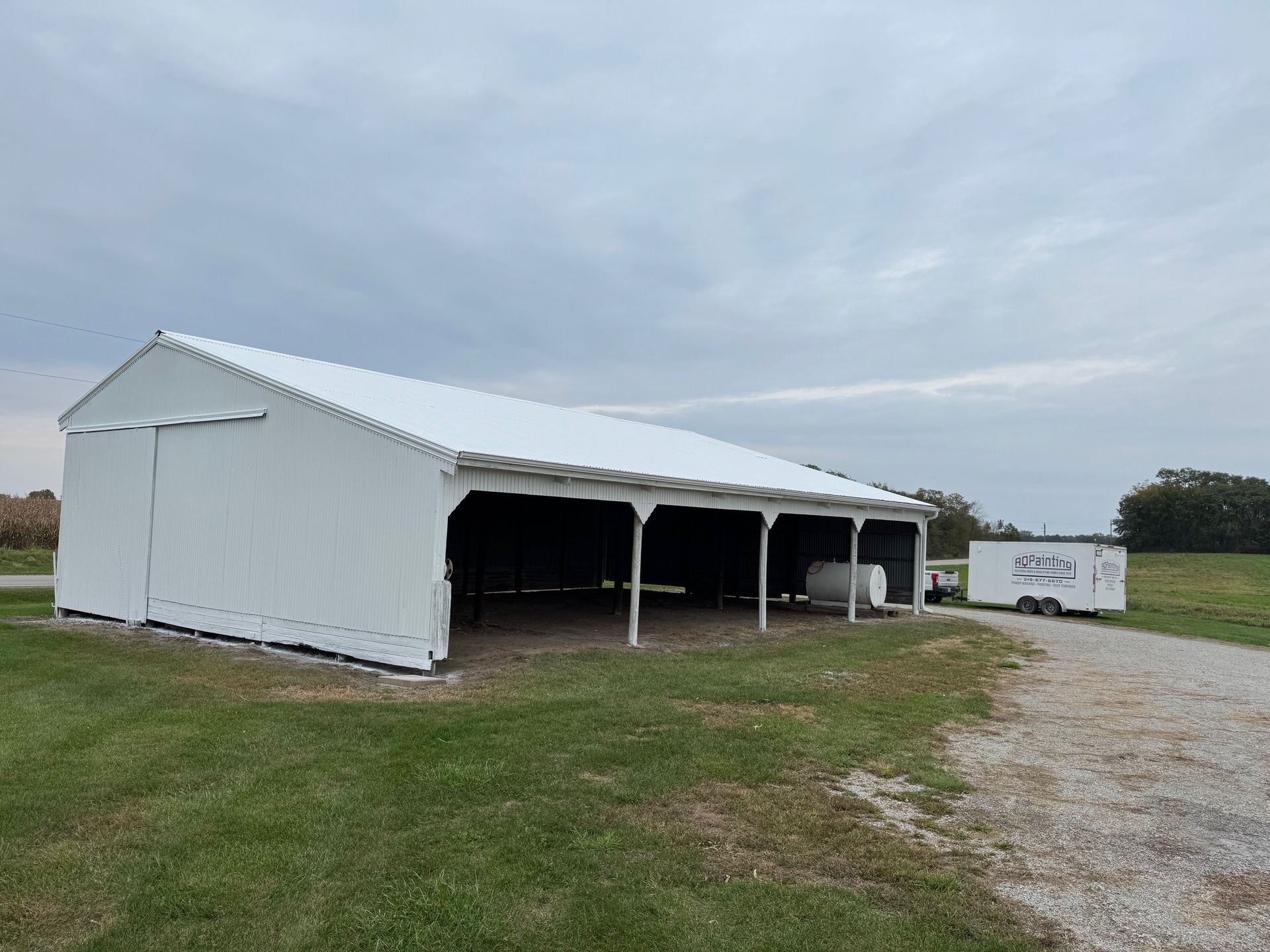 A white, fabric-covered pole barn sits in a grassy field next to a gravel path with a small white trailer parked nearby.