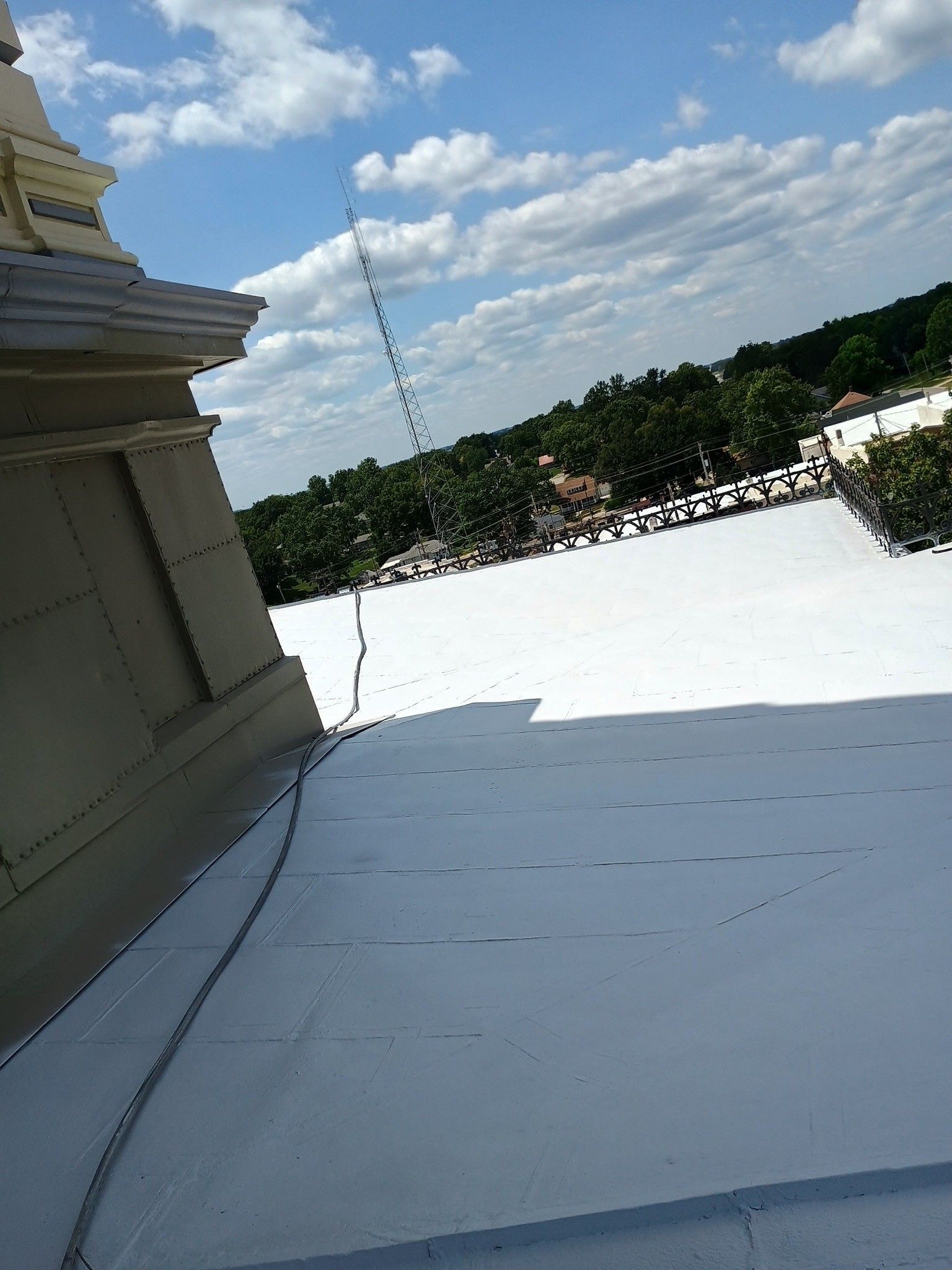 A view from a roof with a white membrane surface, showing a stone structure on the left and trees under a cloudy sky.