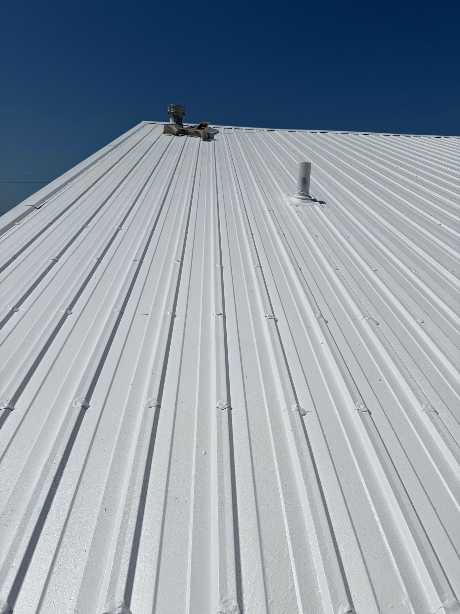 A low-angle view of a white, corrugated metal roof under a clear blue sky, featuring two vent pipes.